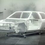 A vintage car frame being painted white in an industrial automotive paint booth.